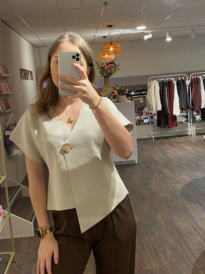 Beige asymmetrische top met V-hals en opvallende gouden knop als detail.
