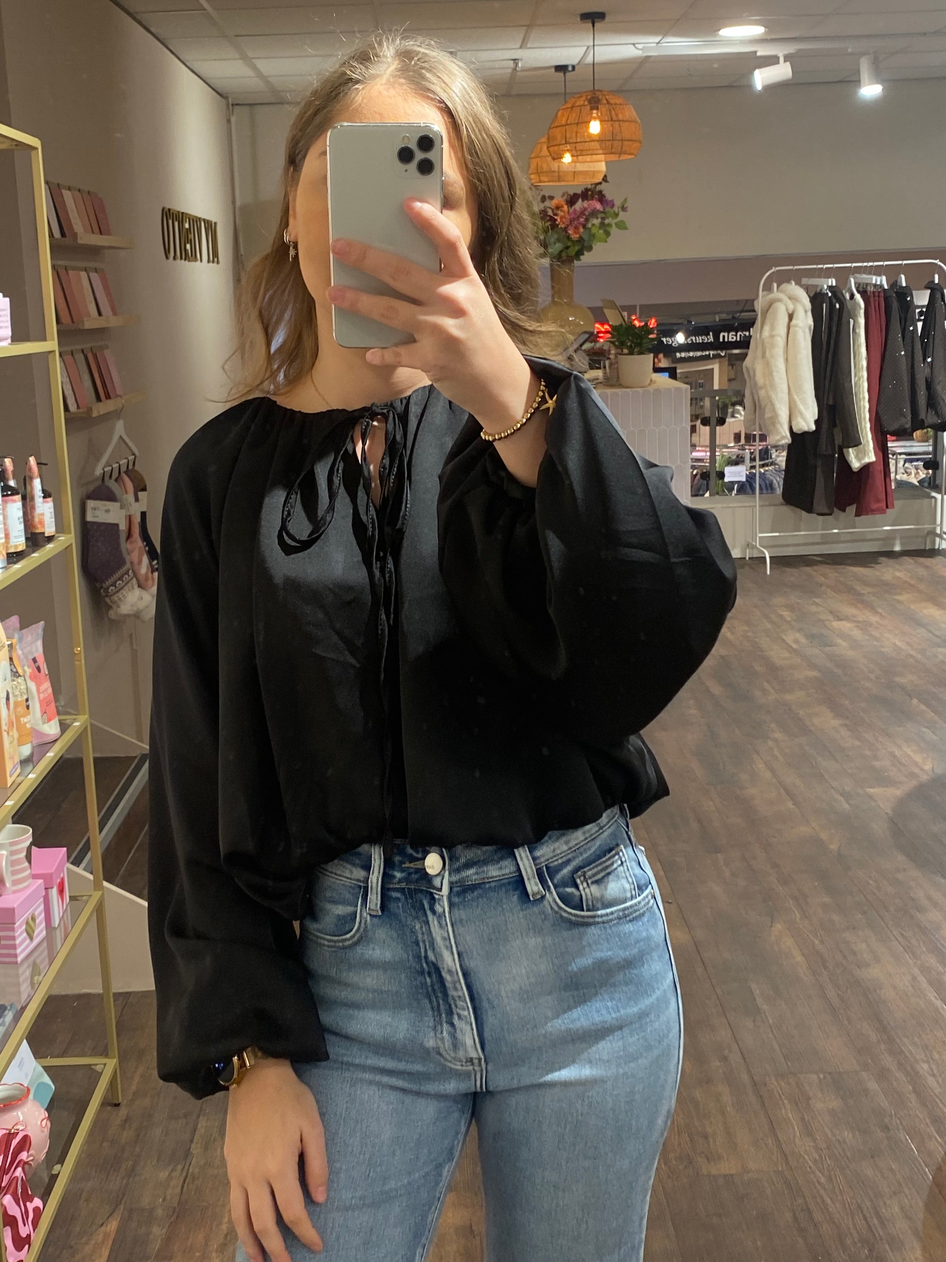 Zwarte satijnachtige blouse met pofmouwen en touwtjes bij de hals.